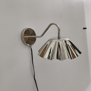Könnte beinhalten: Silberfarbene Wandleuchte mit gerüschtem Lampenschirm. Die Leuchte hat einen kreisförmigen Sockel und einen gebogenen Arm, der den Schirm trägt. Der Schirm hat einen plissierten, gewellten Rand. Ein schwarzes Netzkabel geht von der Basis ab.