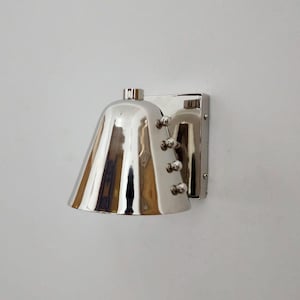 Op de afbeelding: Een gepolijste chromen wandlamp met een klokvormige kap. De lamp heeft een rechthoekige montageplaat en decoratieve klinknagels. De kap reflecteert de omgeving, waardoor een heldere, moderne esthetiek ontstaat. Ontworpen voor binnengebruik.