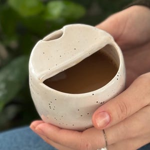 May include: A small, off-white ceramic planter with a unique, rounded shape and a partial opening. The planter is speckled with dark spots and contains a brown substance. The planter is held in a person's hands, with a green plant in the background.