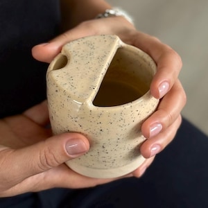 Könnte beinhalten: Ein gesprenkelter Keramikbehälter mit einem einzigartigen Design. Der beige Behälter hat eine gebogene Form mit einer kleinen Öffnung an der Seite und einer größeren Öffnung oben. Der Behälter wird in den Händen einer Person gehalten.