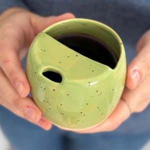Puede incluir: Una taza de cerámica verde claro hecha a mano con un diseño único. La taza tiene una forma redondeada con una sección recortada y un pequeño agujero. El interior está lleno de un líquido oscuro, probablemente café o té. La taza está sostenida por dos manos.