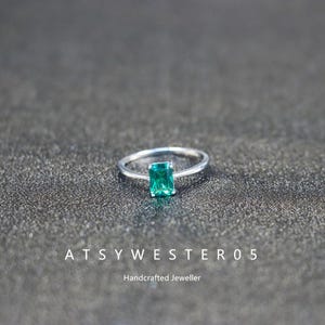 Peut inclure: Une bague en argent avec une pierre précieuse rectangulaire vert émeraude. La bague est posée sur un fond gris foncé. Le texte "ATSYWESTER05" et "Handcrafted Jeweller" sont visibles.