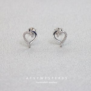 Puede incluir: Un par de pendientes de botón plateados con forma de corazón. Cada pendiente presenta un contorno de corazón con pequeñas piedras transparentes a lo largo de la curva superior. Los pendientes se muestran sobre una superficie azul claro. El texto "ATSYWESTER05 Handcrafted Jewellery" está en la parte inferior.