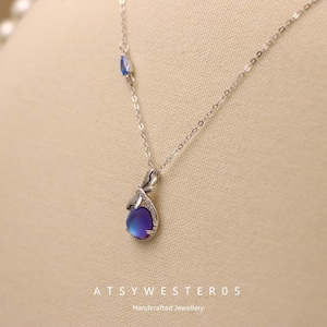 May include: A silver necklace with a teardrop-shaped pendant featuring a blue and purple iridescent stone. The pendant is accented with small clear stones and a delicate silver chain. The image also includes the text "ATSYWESTER05 Handcrafted Jewellery."