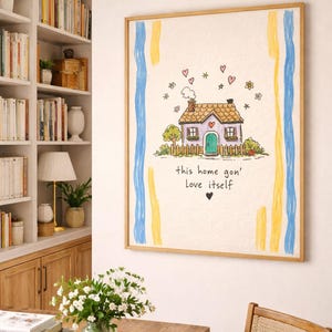 May include: A framed artwork featuring a whimsical house illustration with a purple roof, green door, and a picket fence. The artwork includes the text "this home gon' Love itself" and is decorated with hearts and flowers. Blue and yellow stripes frame the image.