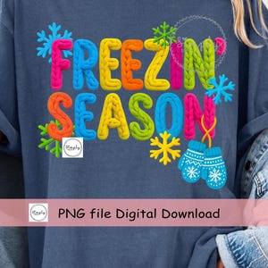 May include: A navy blue t-shirt with the words "FREEZIN' SEASON" in colorful, textured letters. The design includes snowflakes, mittens, and the text "PNG file Digital Download".