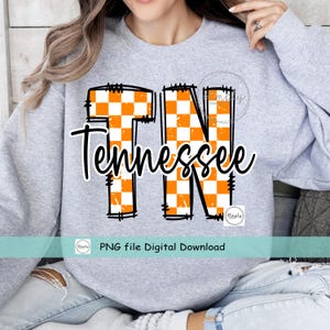 May include: A light gray sweatshirt featuring the word "Tennessee" in a black script font. The letters "TN" are in orange and white checkered pattern. The sweatshirt has a crew neck and long sleeves.