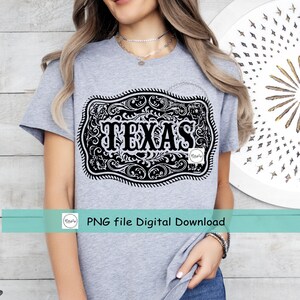 May include: A light gray t-shirt featuring a black and white graphic of a Texas-shaped belt buckle. The buckle design includes ornate scrollwork and the word "TEXAS" in bold letters. The shirt is made of a soft material.