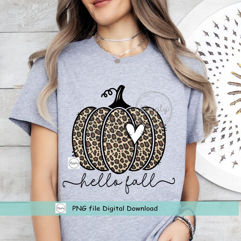 May include: A gray t-shirt with a black and brown leopard print pumpkin with a white heart in the center. The text "hello fall" is written in black cursive below the pumpkin. The text "PNG file Digital Download" is written in black below the text "hello fall".