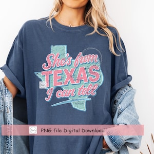 Puede incluir: Camiseta azul marino con un gr&aacute;fico desgastado del contorno del estado de Texas en rosa y turquesa, con el texto "She's from TEXAS I can tell". La camiseta tiene mangas enrolladas.
