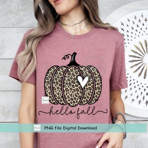 May include: A pink t-shirt with a black and brown leopard print pumpkin with a white heart on it. The text "hello fall" is written below the pumpkin.