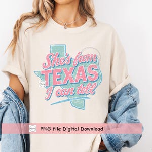 Puede incluir: Camiseta color crema con el texto "She's from TEXAS I can tell" en un estilo retro desgastado. El dise&ntilde;o presenta un contorno de Texas rosa y azul. La camiseta es una descarga digital.