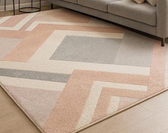 May include: A geometric patterned area rug in shades of pink, gray, and cream. The rug features a modern design with angular shapes and lines. The rug is placed in a living room setting with a gray sofa and decorative pillows.