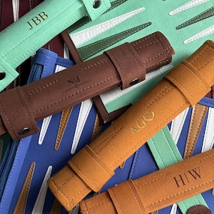 May include: A collection of folded backgammon boards in various colors including mint green, brown, blue, and orange. Each board features a matching leather-like carrying case with a snap closure and personalized initials or symbols embossed.