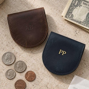 May include: Two semi-circular leather coin pouches, one dark brown with embossed AB initials and one navy blue with gold-stamped PP initials. The pouches are displayed alongside three quarters, two pennies, a one-dollar bill, and a coffee shop receipt.