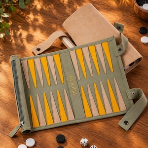 May include: A roll-up backgammon board in olive green fabric with yellow and beige triangular points. The board features the word PHOEBE embossed in the center. Included are white and black circular game pieces and two white dice.