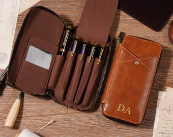 Peut inclure: Une trousse à stylos en cuir marron ouverte, révélant plusieurs stylos avec des détails dorés. Un étui en cuir plus petit, de couleur fauve, avec les initiales "DA" est également visible. Le tout est disposé sur une surface en bois.