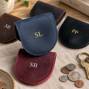 May include: A collection of five horseshoe-shaped leather coin pouches in various colors including blue, burgundy, brown, and black. Each pouch features a unique two-letter monogram embossed on the front, such as SL, SB, or PP, in gold or silver.
