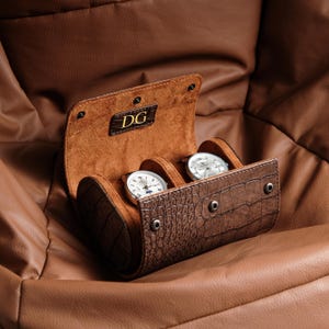 Può includere: Un astuccio porta orologi cilindrico color marrone con texture effetto coccodrillo e chiusura a bottoni automatici, dotato di rivestimento interno color cuoio e lettere DG impresse sulla patta. Contiene due orologi argentati a quadrante bianco.