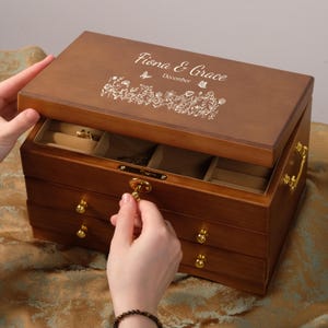 Pode incluir: Uma caixa de joias de madeira com tampa articulada, com o texto gravado "Fiona & Grace December" e desenhos florais. A caixa tem três gavetas com puxadores dourados e um fecho dourado. A tampa está sendo aberta para revelar o interior.