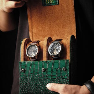 Può includere: Un astuccio verde con texture a coccodrillo che contiene due orologi. L'astuccio è aperto, rivelando un interno marrone. Gli orologi hanno cinturini in pelle nera e quadranti argentati e dorati. L'astuccio ha le iniziali "DM" su un'etichetta verde.