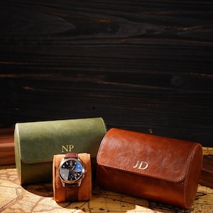 May include: A brown leather watch with a black face and a brown leather strap is displayed on a tan watch pillow. Two leather watch cases, one olive green with "NP" and one brown with "JD", are also shown.