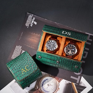 Peut inclure: Plusieurs montres de luxe et étuis. Deux montres avec des bracelets en cuir marron sont présentées dans un étui vert texturé façon crocodile. Un petit étui et une troisième montre sont également visibles. Les étuis sont estampés d'initiales.