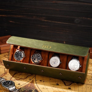 以下が含まれることがあります： 4つの時計が入った、開いた緑色の革製時計ケース。ケースにはフラップクロージャーがあり、上部にはイニシャル「HD」がエンボス加工されています。銀色の時計が地図の上に置かれています。