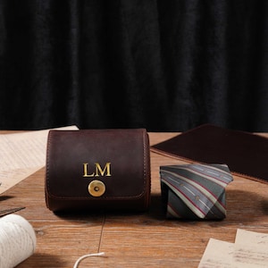 Può includere: Un astuccio per orologio in pelle marrone scuro con le iniziali dorate "LM" e una chiusura a scatto dorata. Accanto, una cravatta a righe piegata con dettagli rossi e bianchi. Gli oggetti sono su una superficie di legno.