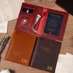 May include: A collection of leather travel wallets in various shades of brown and red, displayed on a wooden surface. One wallet is open, revealing a passport and cash. Two other wallets are closed, with gold initials. The open wallet has a zipper and a small pocket.