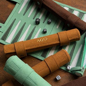 Puede incluir: Un tablero de backgammon verde menta con puntas blancas y verde oscuro, acompañado de varios estuches de ante enrollados en marrón, tostado y verde menta. Un estuche tostado presenta el texto dorado AGO con un corazón, y hay dados negros.