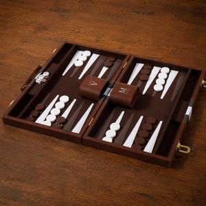 May include: A wooden backgammon set featuring a dark brown board with white and brown triangular points. The set includes white and brown checkers, two dice, and two brown leather cups embossed with the letters A and M in gold foil.