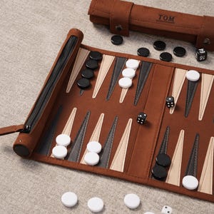 May include: A brown backgammon set with black and white game pieces. The board is open, revealing the playing surface with triangular points. A zippered case and dice are also present. The word "TOM" is printed on the case.
