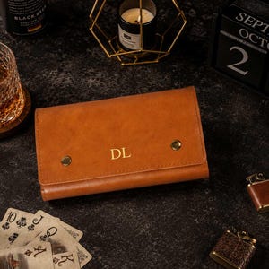 May include: A rectangular tan leather wallet featuring a fold-over flap secured by two gold-toned metal snaps. The center of the flap is embossed with the gold-colored initials DL. The wallet has a smooth, matte finish and a structured design.
