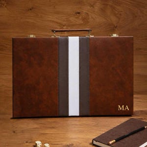 May include: A rectangular brown briefcase with a central vertical stripe featuring a white band flanked by two dark brown strips. The briefcase has a brown handle, gold-toned metal latches, and the gold-embossed letters MA in the bottom right corner.