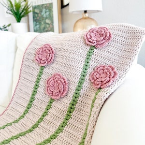 May include: A crocheted throw blanket with a light pink background and green stripes. The blanket features four pink crocheted flowers.