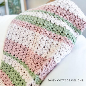 Puede incluir: Una manta de ganchillo con rayas de color rosa, verde y crema. La manta está doblada y drapeada sobre una superficie blanca. El texto "DAISY COTTAGE DESIGNS" es visible en la esquina inferior derecha de la imagen.