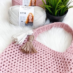 May include: A pink crocheted tote bag with two tassels. The bag is made with a chunky yarn and has a simple, open weave. Two spools of yarn are visible in the background, one pink and one white.