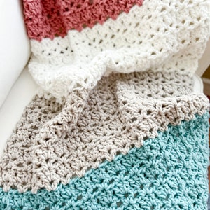 Pu&ograve; includere: Una coperta all'uncinetto con quattro strisce di diversi colori. Le strisce sono turchese, beige, bianco e rosa. La coperta &egrave; piegata e drappeggiata su un divano bianco.