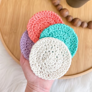 Puede incluir: Cuatro almohadillas de desmaquillaje de ganchillo en diferentes colores: coral, turquesa, lavanda y blanco. Las almohadillas están apiladas una encima de la otra y se sostienen en una mano.