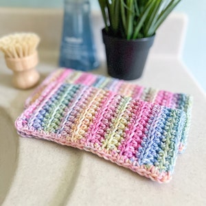 May include: Two rainbow colored crochet dishcloths, made with a textured stitch, are sitting on a white countertop.