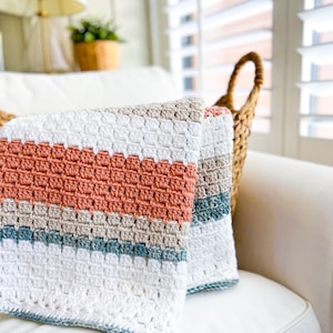May include: A crocheted throw blanket with stripes of white, beige, light blue, and peach. The blanket is folded and placed on a white sofa.