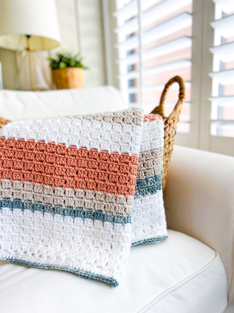 May include: A crocheted throw blanket with stripes of white, beige, light blue, and peach. The blanket is folded and placed on a white sofa.