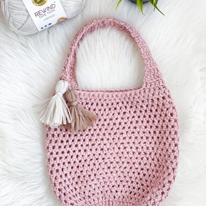 May include: A pink crocheted tote bag with two tassels. The bag is made with a simple stitch pattern and has a wide, open top. The bag is sitting on a white fluffy surface.
