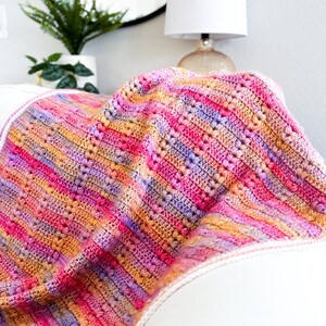 May include: A multi-colored crocheted blanket with a striped pattern in shades of pink, orange, and purple. The blanket is folded and draped over a white couch.