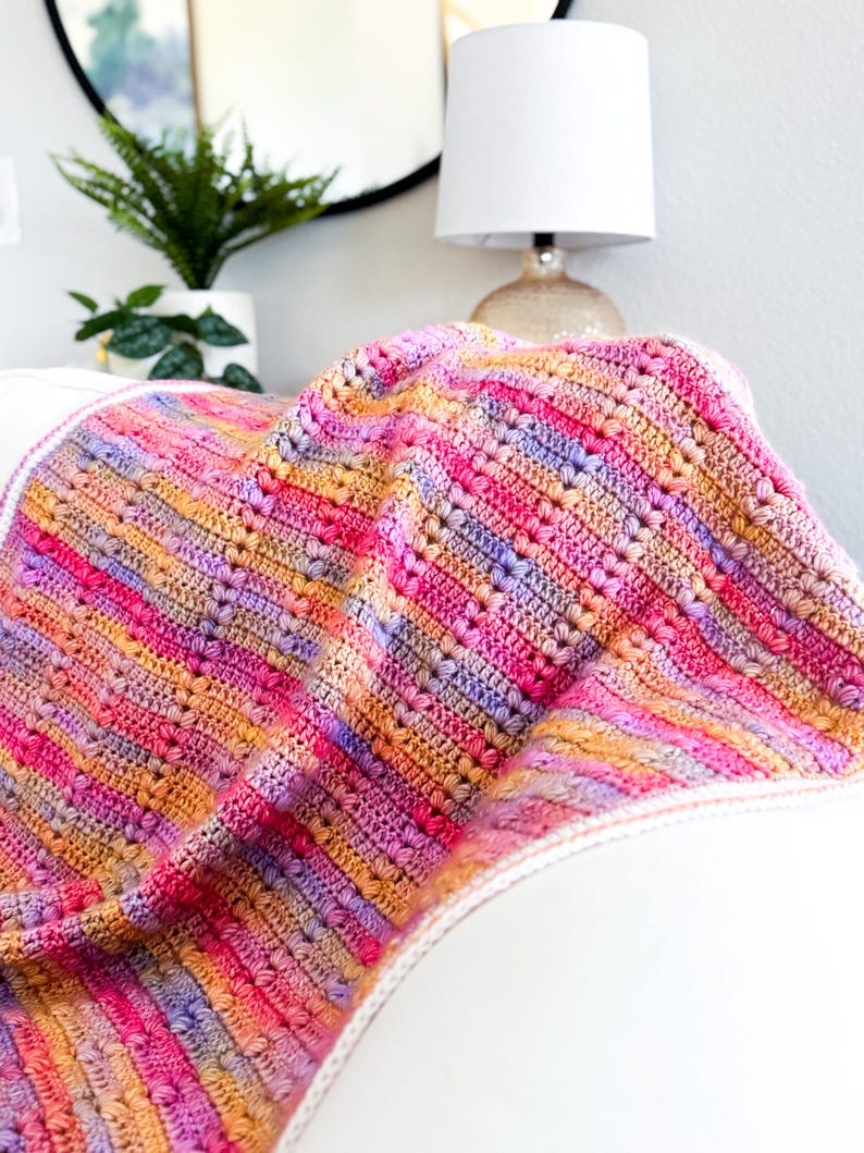 May include: A multi-colored crocheted blanket with a striped pattern in shades of pink, orange, and purple. The blanket is folded and draped over a white couch.
