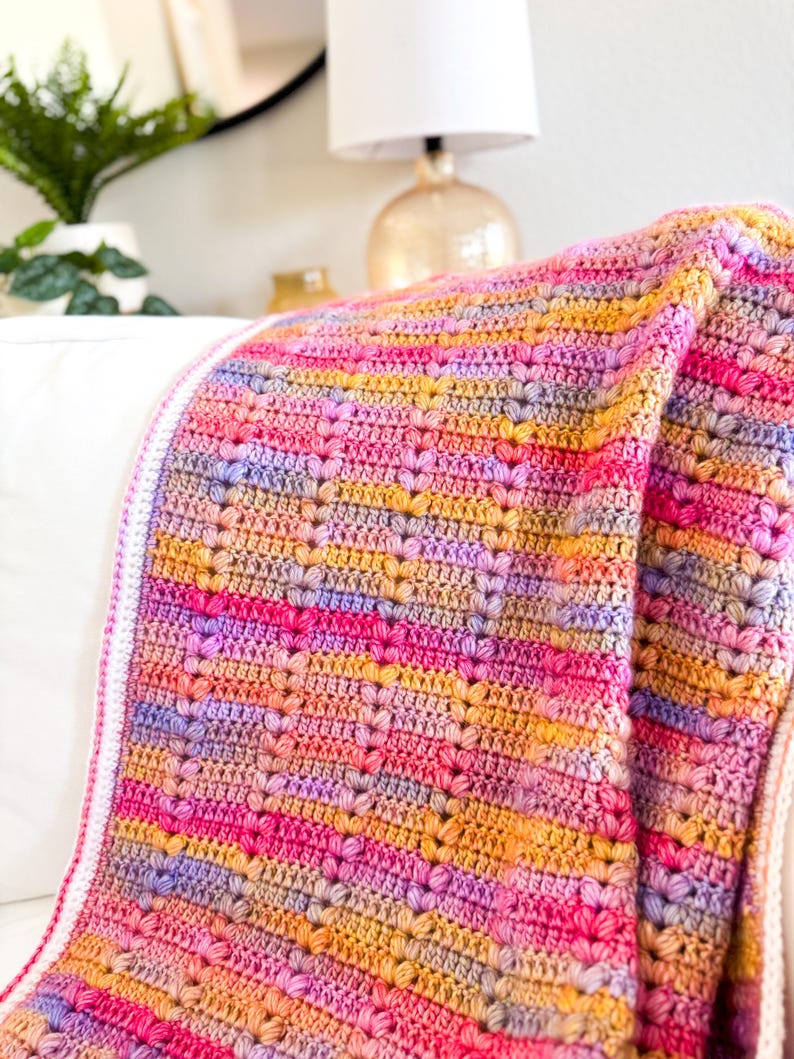 May include: A colorful crocheted blanket with a heart stitch pattern in shades of pink, purple, orange, and yellow. The blanket is folded and draped over a white couch.