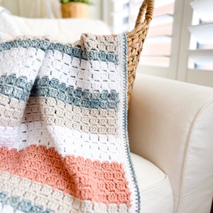 May include: A crocheted throw blanket with stripes of white, beige, light blue, and peach. The blanket is draped over a white sofa.