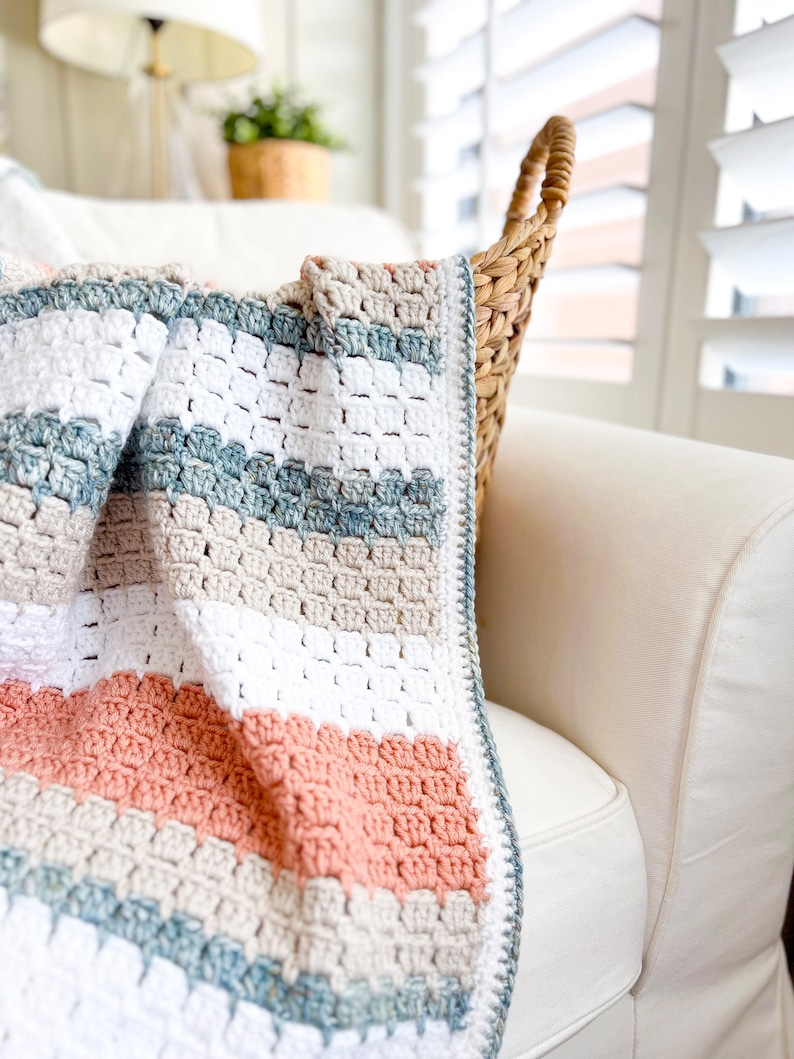 May include: A crocheted throw blanket with stripes of white, beige, light blue, and peach. The blanket is draped over a white sofa.
