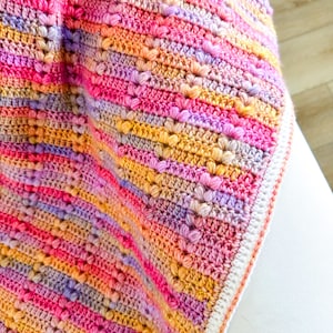 May include: A multi-colored crocheted baby blanket with a white border. The blanket is made with a textured stitch that creates a raised pattern. The colors are pink, purple, orange, and yellow.